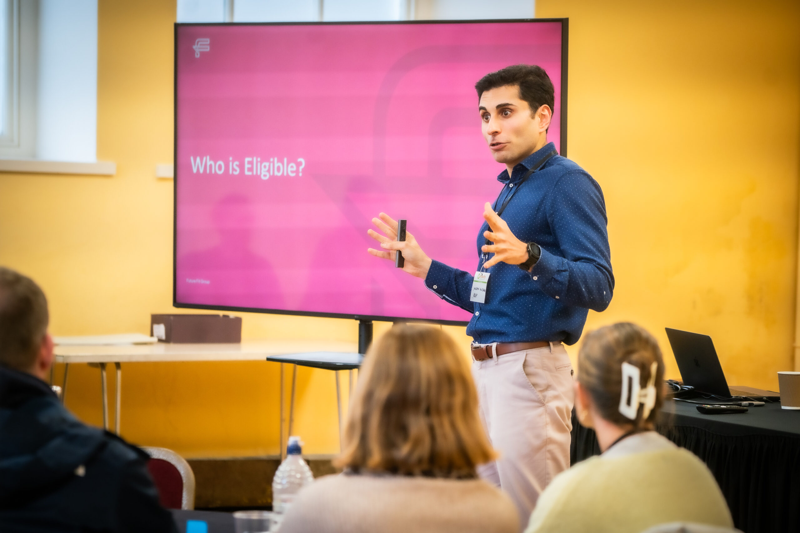 Man stood in front of a screen with writing 'Who is eligable', presenting to an audience.